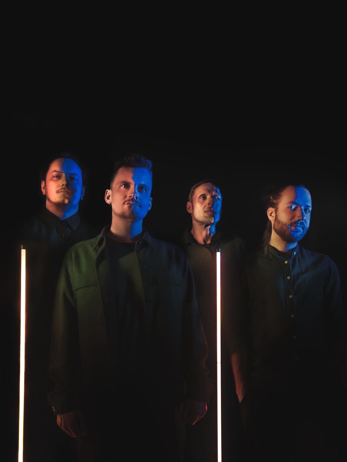 DRIVEN BY CLOCKWORK — the four members side by side between two neon tubes, lit blue and amber.